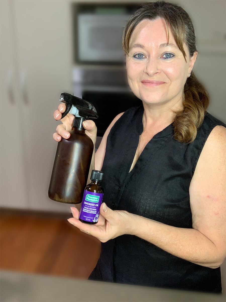 Image description: woman standing in a kitchen holding a dark glass spray bottle and small bottle of lavender oil